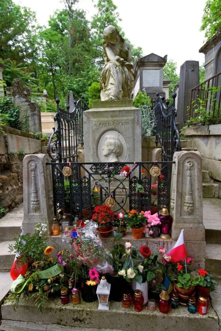 The tomb of Frédéric Chopin at Père Lachaise Cemetery is adorned with a sorrowful marble sculpture, an engraved portrait of the composer, and an array of flowers, candles, and tributes, including a Polish flag left by admirers.