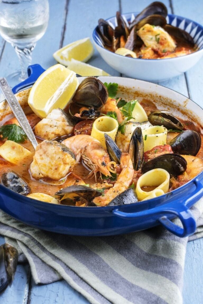 A vibrant seafood stew with shrimp, mussels, fish, and pasta in a rich tomato broth, served in a blue dish with lemon wedges—perfect for a Marseille Food Guide.