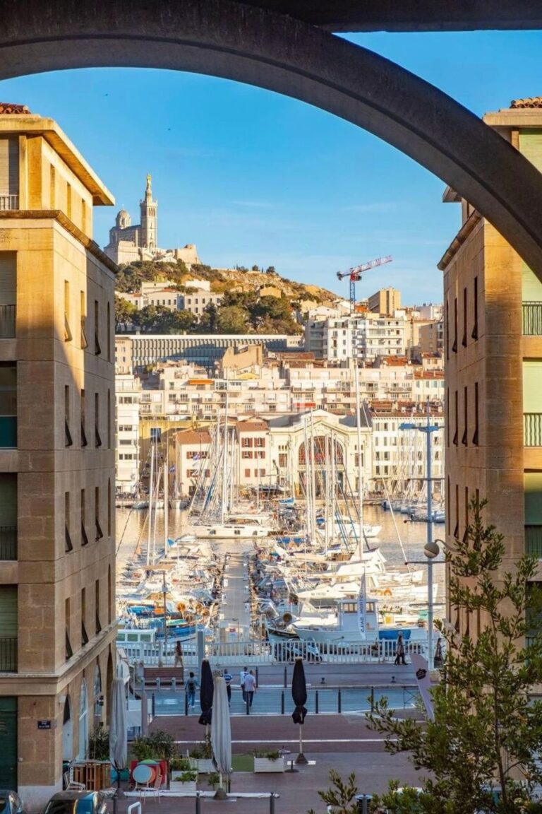 A scenic view of Marseille’s Old Port, framed by buildings, with sailboats docked in the harbor and the Notre-Dame de la Garde basilica atop a hill—one of the top things to do in Marseille France.