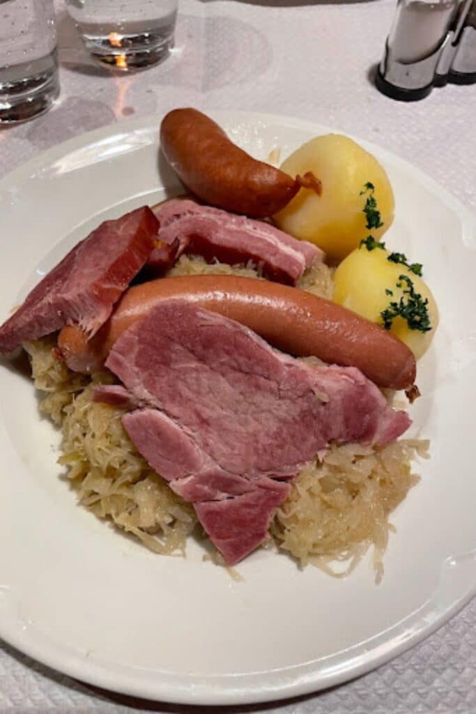 Plate of traditional Alsatian choucroute garnie with sauerkraut, sausages, smoked meats, and boiled potatoes, served at a restaurant in Strasbourg. This hearty regional dish is a delicious way to experience local cuisine during one day in Strasbourg. The meal is beautifully plated with simple garnishes.