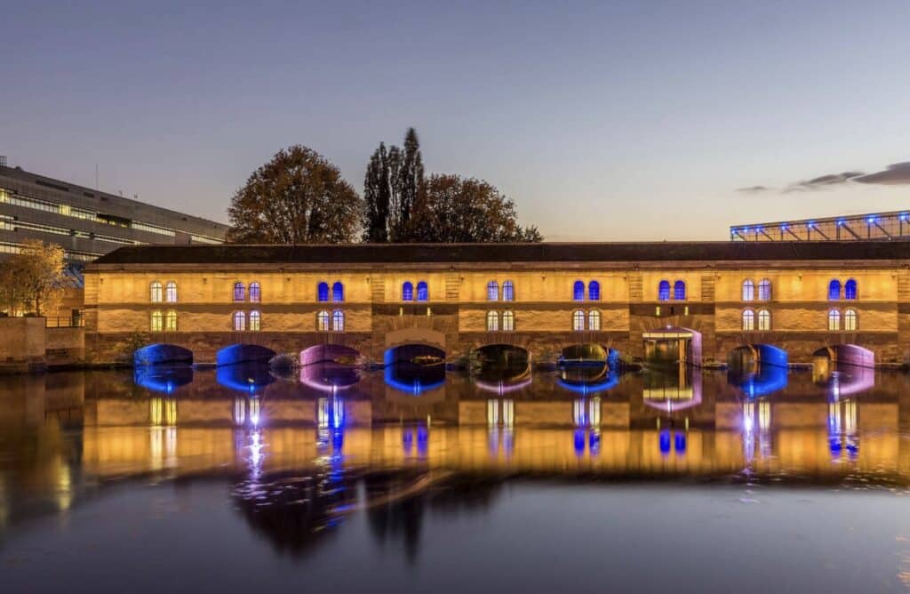 The Barrage Vauban in Strasbourg lit up at night with colorful blue and yellow lights reflecting on the river. This historic stone bridge and dam is a must-see landmark for anyone spending one day in Strasbourg. Modern buildings and autumn trees frame the scene in the background.