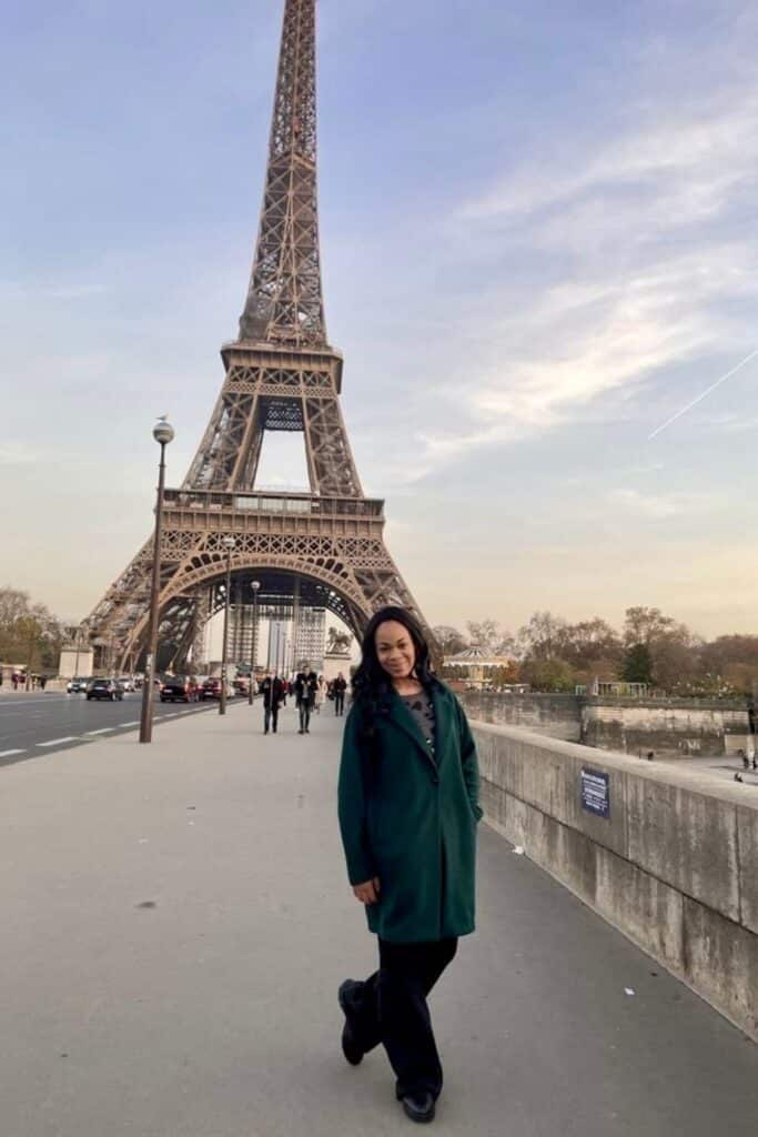 A woman in a dark green coat strolls along the Seine River near the Eiffel Tower at golden hour, with a soft pastel sky overhead and light foot traffic behind her. Her relaxed smile and confident pose capture the dreamy, bucket-list moment of standing before Paris’s most iconic landmark during one day in Paris.