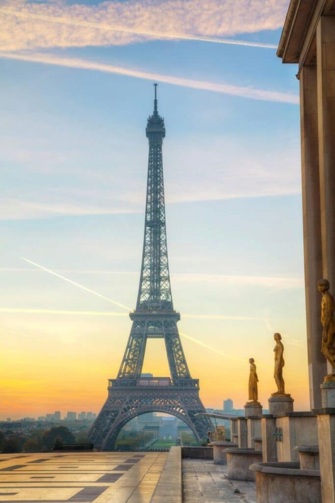 The Eiffel Tower stands tall in the soft morning light, viewed from the Trocadéro esplanade at sunrise. The golden sky glows behind delicate contrails and bronze statues, creating a serene, picture-perfect moment that captures the quiet beauty of one day in Paris before the city wakes.