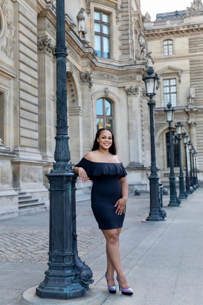 A Paris travel blogger poses with a smile, leaning against an ornate black lamppost in the Louvre courtyard, surrounded by the museum’s grand historic architecture, intricate stone carvings, and a row of elegant lampposts.