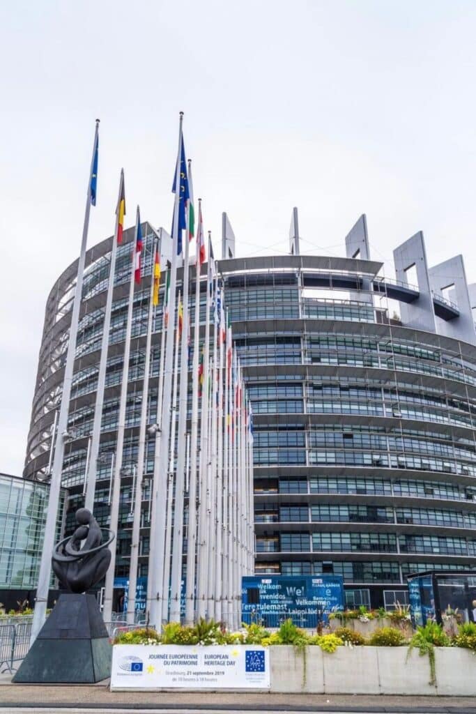 Modern glass facade of the European Parliament building in Strasbourg, with rows of European Union member flags and a sculpture in front. The curved architecture and tall flagpoles make it one of the city’s most recognizable landmarks. Visiting this important site is a unique stop during one day in Strasbourg.