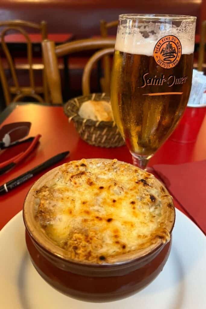 A rich bowl of French onion soup topped with bubbly, golden-brown cheese sits on a red café table beside a cold glass of Saint-Omer beer. With rustic wooden chairs, a bread basket, and classic bistro ambiance in the background, this hearty meal captures the comforting culinary experience of one day in Paris.