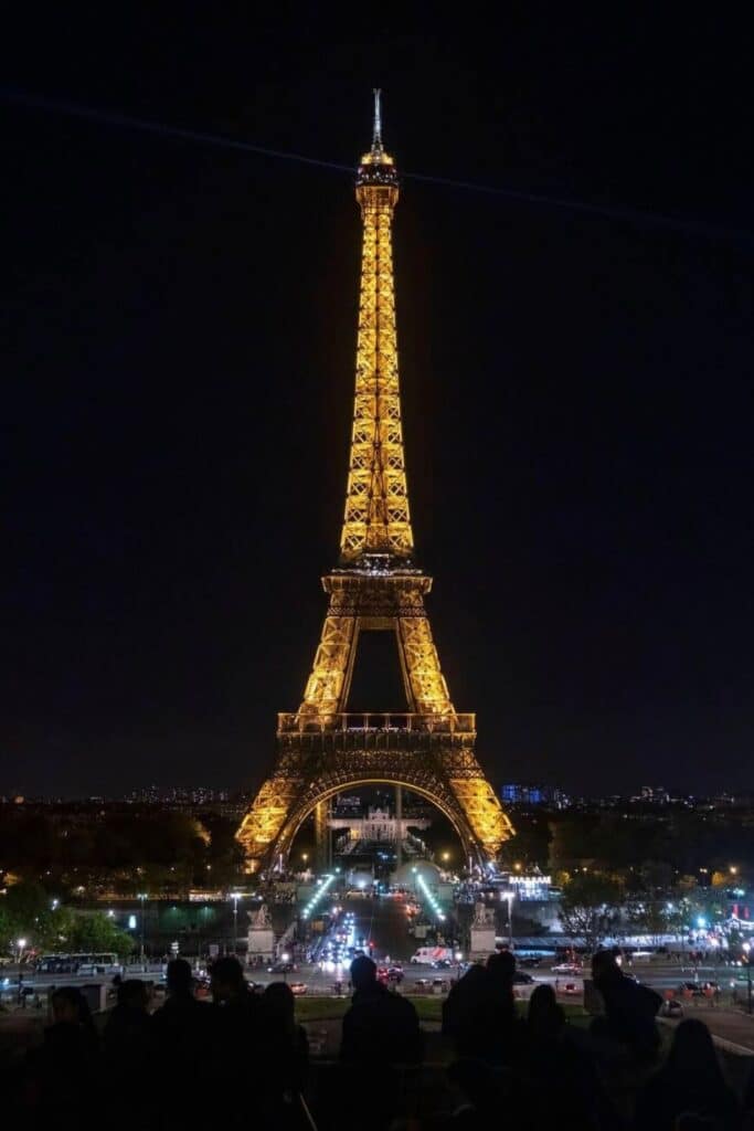 The Eiffel Tower glows with golden lights against the dark Paris sky, as viewed from the Trocadéro at night. Crowds gather in silhouette below, admiring the illuminated landmark and the bustling city beyond—a dazzling finale to one day in Paris.