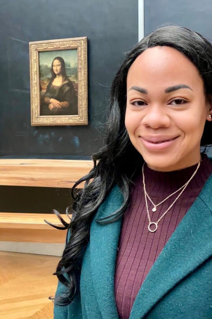 A smiling woman poses in front of Leonardo da Vinci’s Mona Lisa at the Louvre Museum, with the famous portrait framed behind protective glass in the background. Her expression reflects the excitement of seeing one of the world’s most iconic artworks up close—a cultural highlight during one day in Paris.