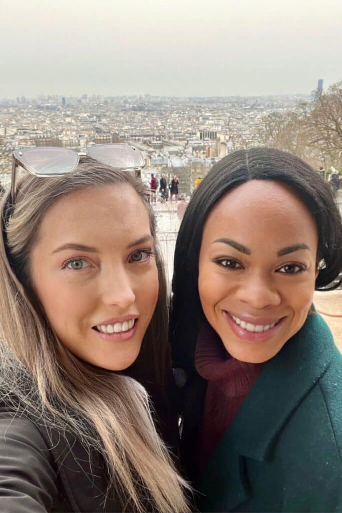 Two smiling women take a selfie at the top of Montmartre, with a panoramic view of the Paris skyline stretching out behind them. Surrounded by soft winter light and the rooftops of the city, this joyful moment captures the magic of discovering Paris with a friend during one day in Paris.