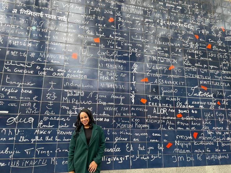 A woman in a green coat smiles in front of the Mur des Je t’aime (I Love You Wall) in Montmartre, a deep blue tiled mural featuring “I love you” written in over 250 languages. This romantic and colorful landmark is a heartwarming stop for travelers exploring art, culture, and connection during one day in Paris.