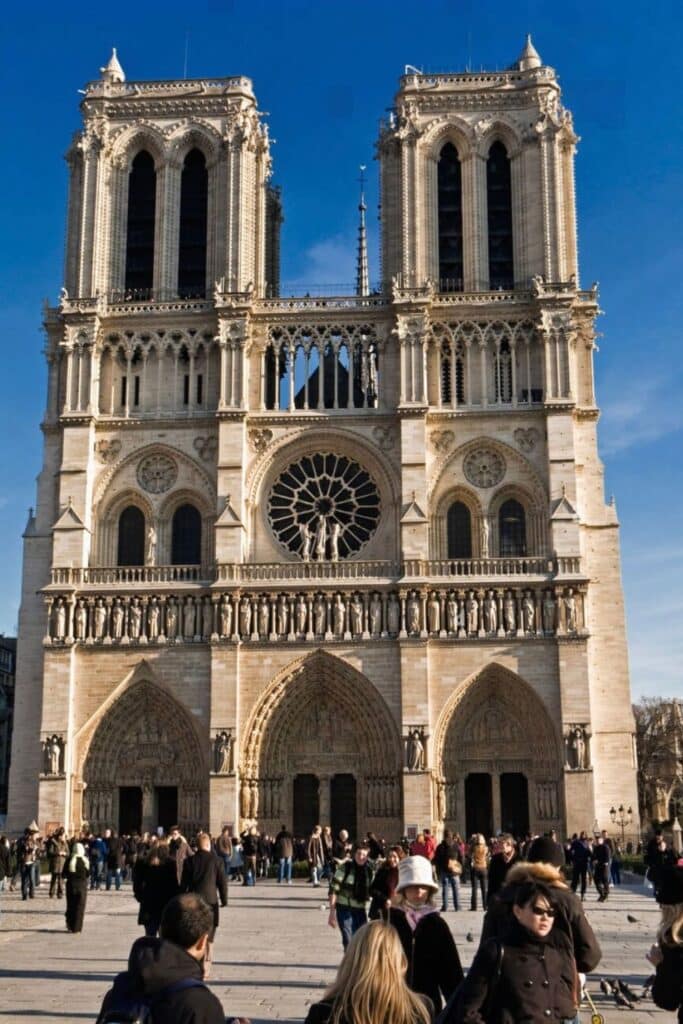 The magnificent west façade of Notre-Dame Cathedral stands tall against a clear blue sky, with intricate stone carvings, twin bell towers, and its famous rose window on full display. Tourists gather in the bustling square below, admiring this architectural masterpiece during one day in Paris.
