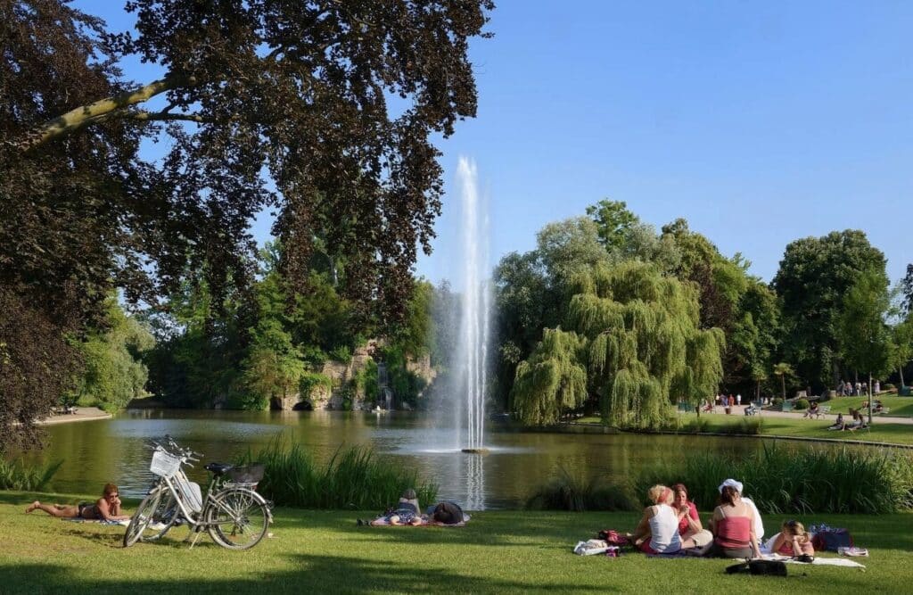 People relaxing and having a picnic on the grass near a large fountain and pond in Parc de l'Orangerie, Strasbourg, on a sunny day. Bicycles are parked nearby, and lush green trees surround the water. Visiting this peaceful park is a great way to unwind during one day in Strasbourg.