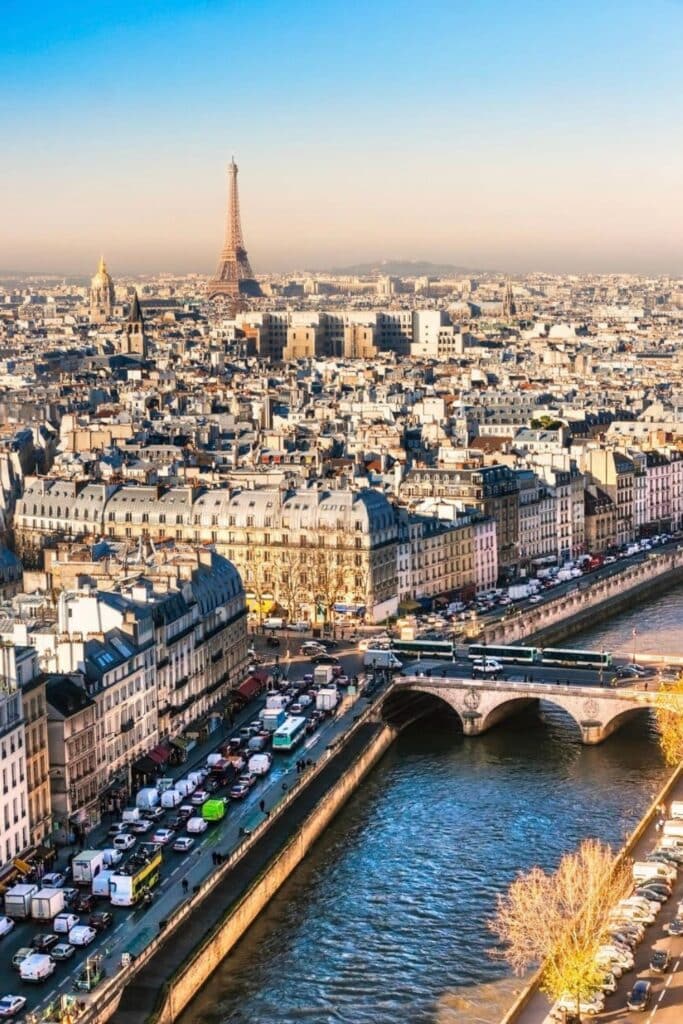 A sweeping aerial view of Paris reveals a sea of cream-colored rooftops, the winding Seine River, and the Eiffel Tower rising in the distance under a soft blue sky. With Haussmannian buildings, charming bridges, and lively traffic below, this iconic cityscape captures the vibrant energy of one day in Paris.