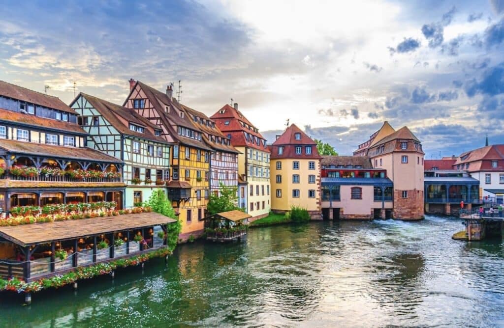 Colorful half-timbered houses and waterfront restaurants line the scenic canals of Petite France in Strasbourg, with flowers and reflections in the water. This charming medieval district is one of the top places to visit during one day in Strasbourg. The sky is partly cloudy, adding to the fairytale atmosphere of the historic neighborhood.
