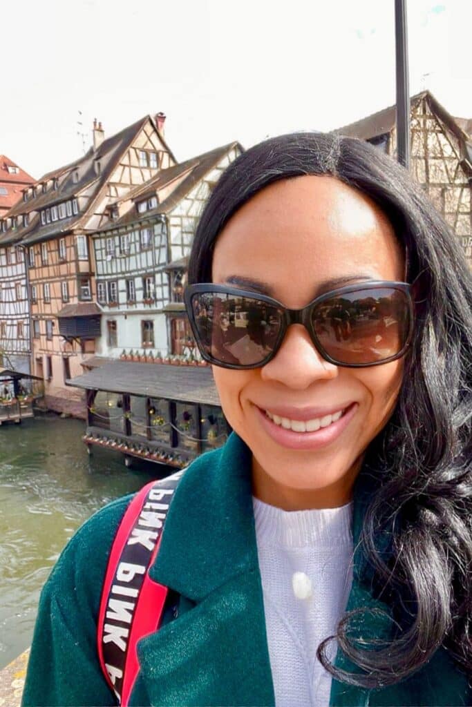 Woman taking a selfie in Petite France, Strasbourg, with half-timbered houses and a canal in the background on a bright day. This charming medieval district is a must-see for anyone spending one day in Strasbourg. The woman is smiling, wearing sunglasses and a green coat, capturing a perfect travel moment.