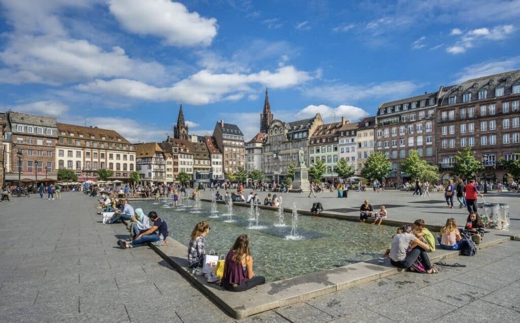 Place Kléber, the main square in Strasbourg, filled with people sitting around a modern fountain on a sunny day. Historic buildings with colorful facades and Strasbourg Cathedral's spire are visible in the background. This lively square is a popular spot to relax and people-watch during one day in Strasbourg.
