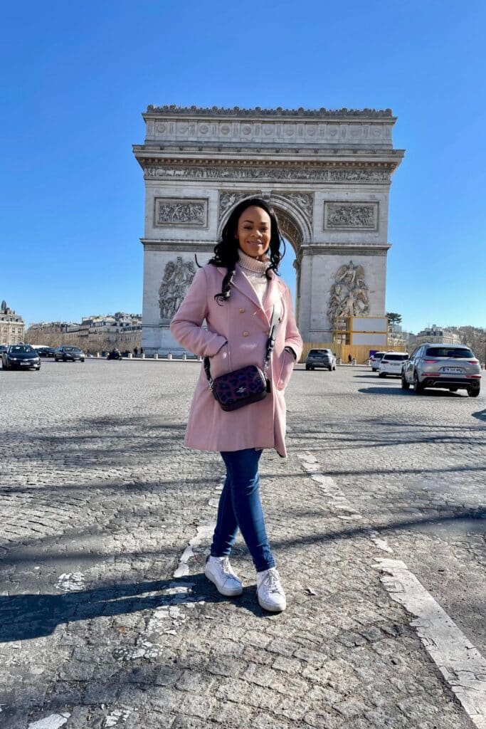 A stylish woman in a blush pink coat poses confidently in front of the Arc de Triomphe on a crisp, sunny day in Paris. The monument’s neoclassical details stand out against a bright blue sky, marking a classic sightseeing stop during one day in Paris.