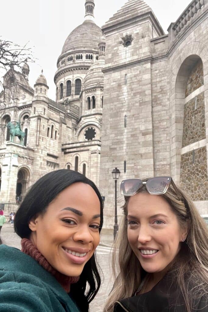 Two smiling women pose for a selfie in front of the Sacré-Cœur Basilica in Montmartre, Paris, with the intricate domes of the church rising behind them—capturing a joyful moment during one day in Paris exploring its artistic and historic neighborhoods.