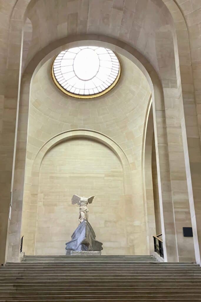 The Winged Victory of Samothrace statue displayed atop a grand staircase under a domed skylight at the Louvre Museum in Paris—one of the must-see highlights during one day in Paris for art and history lovers.
