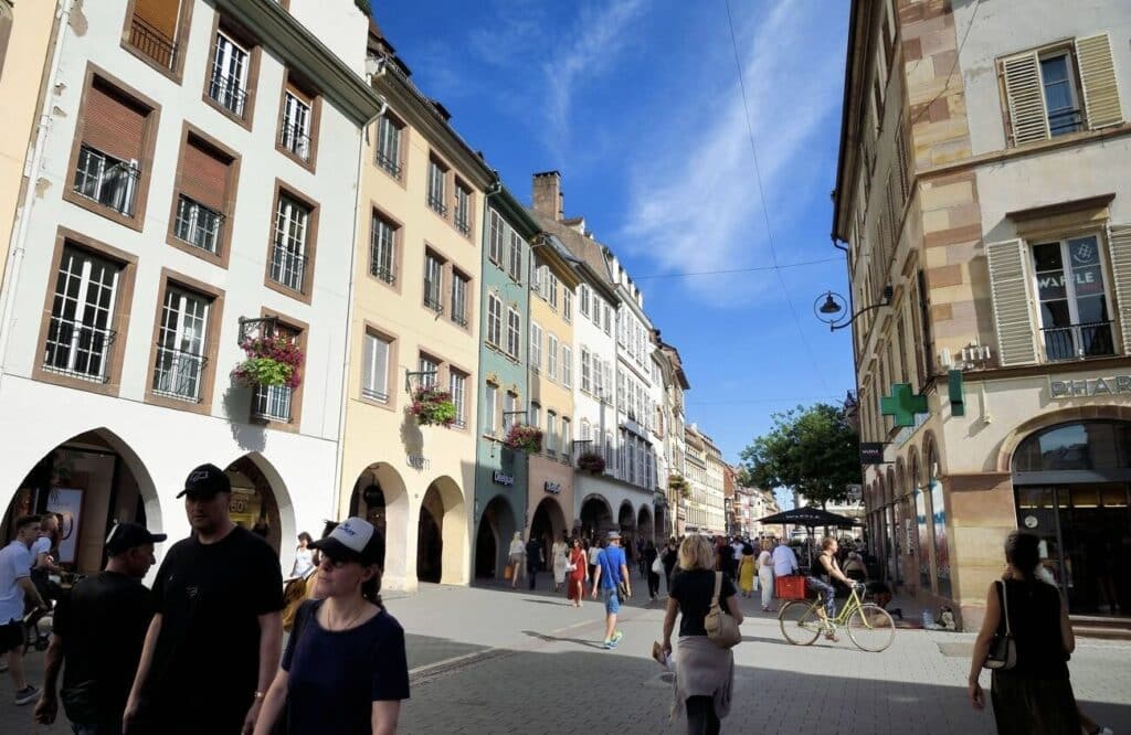 Rue des Grandes Arcades, a busy shopping street in Strasbourg, filled with people walking and biking on a sunny day. The street is lined with colorful historic buildings featuring arched arcades and flower-filled balconies. This lively pedestrian area is a great place to explore during one day in Strasbourg for shopping and local atmosphere.