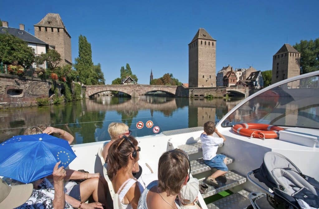 Tourists on a Batorama boat cruise in Strasbourg, passing by the historic Ponts Couverts and medieval towers on a sunny day. The calm river reflects the stone bridges and towers, creating a picturesque scene. A boat tour like this is a great way to explore the city’s canals during one day in Strasbourg.