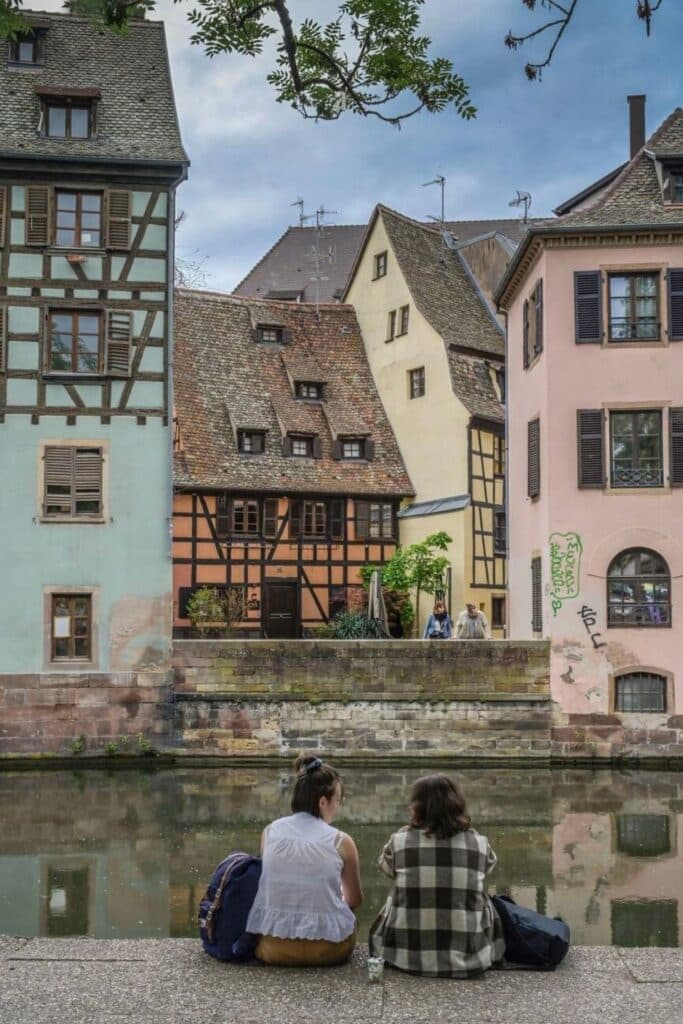 Two people sitting by a peaceful canal in Petite France, Strasbourg, with colorful half-timbered houses and pastel buildings reflected in the water. The scene captures the charming medieval architecture and relaxed atmosphere visitors can enjoy during one day in Strasbourg. A tree branch frames the view above, adding to the cozy riverside setting.
