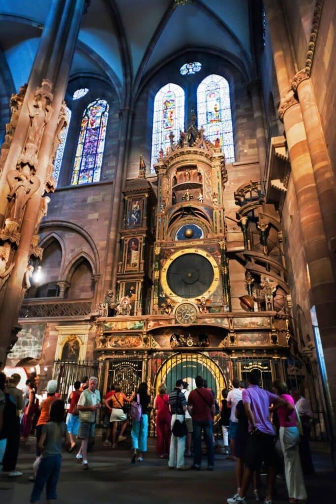 Interior view of Strasbourg Cathedral showing the famous Astronomical Clock, a richly decorated timepiece with golden details, figurines, and zodiac signs. Tourists gather to admire the clock beneath tall stained glass windows. Visiting this unique masterpiece is one of the top highlights during one day in Strasbourg.