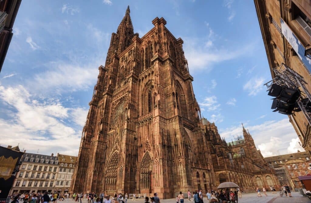 Front view of Strasbourg Cathedral (Notre-Dame), a towering Gothic masterpiece with intricate stone carvings and a single spire, under a partly cloudy sky. Tourists gather in the square below, admiring the cathedral’s stunning facade. Visiting this iconic landmark is a must during one day in Strasbourg.