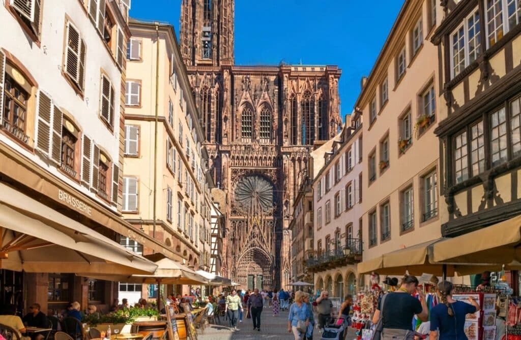 Bustling view of Rue Mercière in Strasbourg with cafes, market stalls, and people walking toward the towering Strasbourg Cathedral under a bright blue sky. The Gothic facade of the cathedral dominates the background, framed by historic buildings. This iconic street is a must-visit during one day in Strasbourg for shopping, dining, and sightseeing.