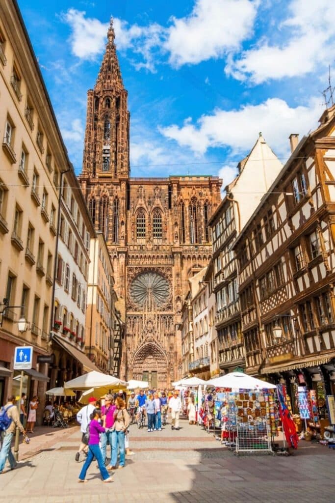 Bustling pedestrian street in Strasbourg lined with half-timbered houses and market stalls, leading to the majestic Strasbourg Cathedral under a bright blue sky. People are walking and browsing souvenirs along the lively street. This iconic view is a highlight for anyone spending one day in Strasbourg.