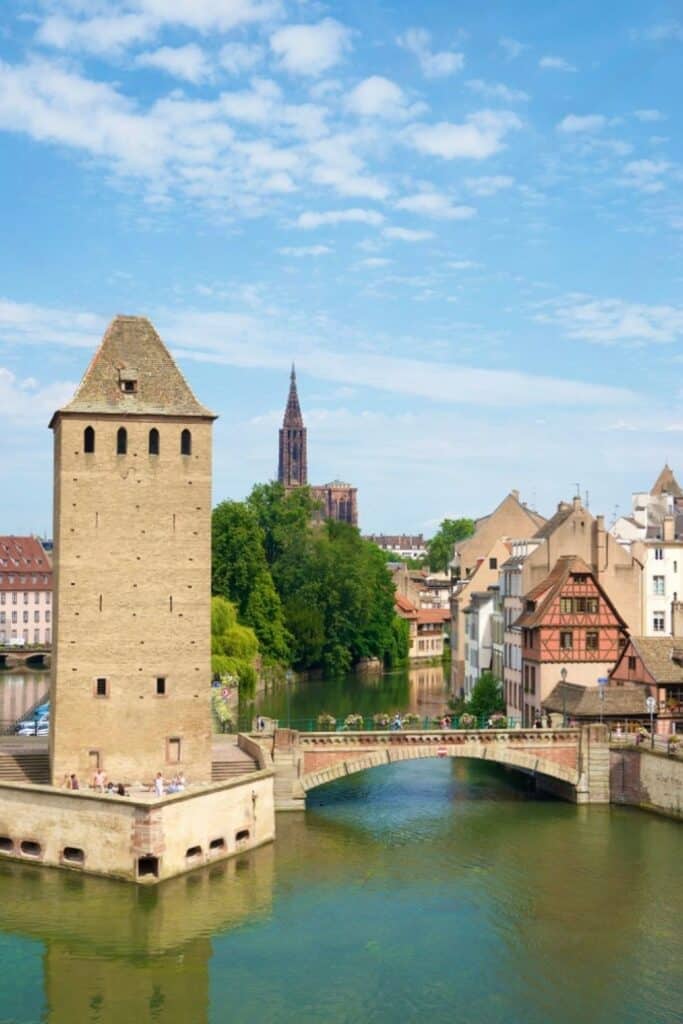 View of the Ponts Couverts towers and bridge in Strasbourg, with the cathedral spire rising in the background and colorful historic houses along the canal. The calm water reflects the medieval architecture and green trees. This scenic spot is a must-see during one day in Strasbourg.