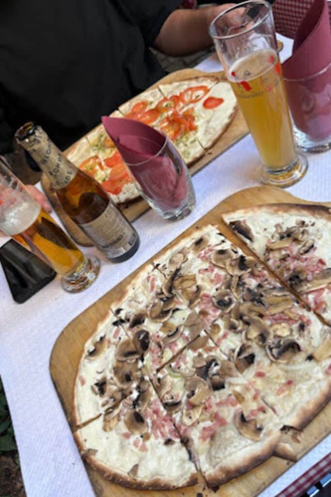 Traditional Alsatian tarte flambée (flammekueche) topped with mushrooms, bacon, and cream, served on wooden boards at a restaurant in Strasbourg. Glasses of beer and other variations of the dish are also on the table. Tarte flambée is a must-try local specialty during one day in Strasbourg.