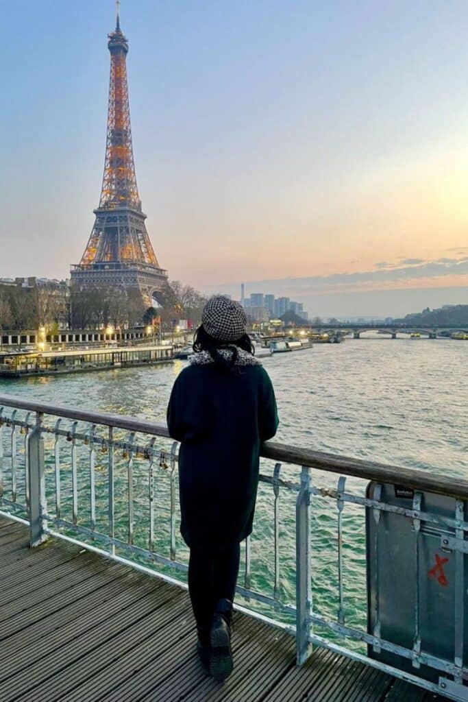 A woman in a houndstooth beret and dark coat stands on a pedestrian bridge at sunset, gazing at the Eiffel Tower glowing with lights—an unforgettable moment during one day in Paris.