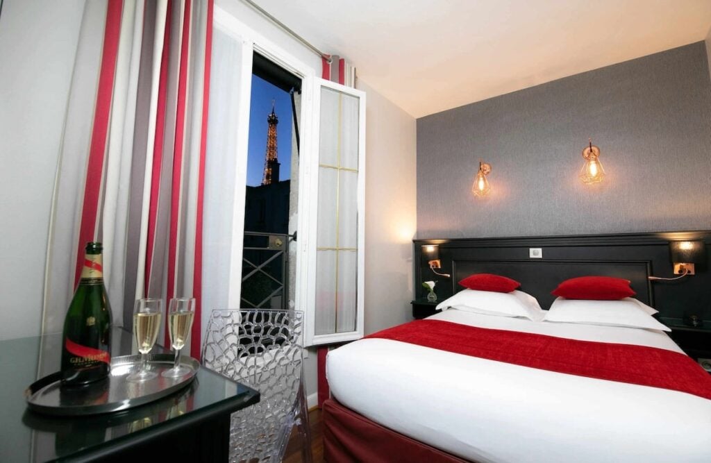 Cozy room at Hôtel Eiffel Rive Gauche with red accents, a neatly made bed, and an open window showcasing the Eiffel Tower illuminated at night, perfect for cheap hotels with Eiffel Tower view.