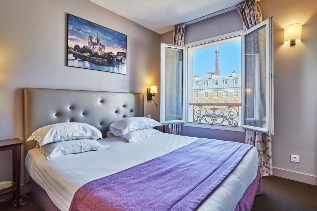 Cozy room at Hotel Le Relais Saint Charles with soft purple accents, a large bed, and an open window showcasing a charming view of the Eiffel Tower, perfect for cheap hotels with Eiffel Tower view.